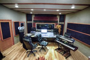 Wide angle shot of a modern high tech recording studio that includes various methods of recording, digital and analog, a wooden floor, sound console, board or desk, computer workstation, electronic keyboards and a window into an isolation booth.